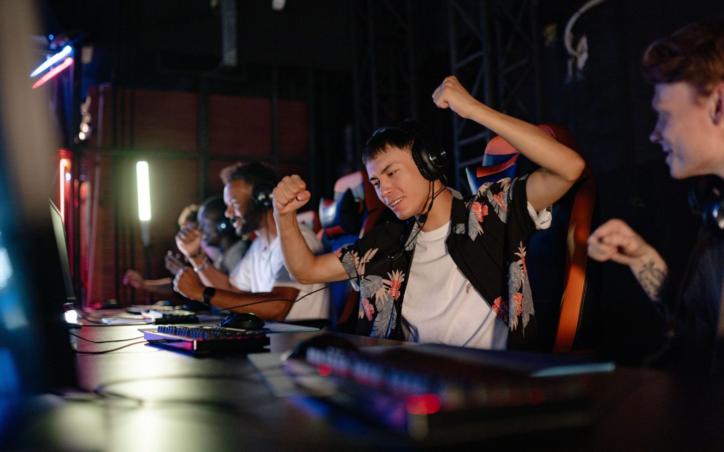 Des joueurs souriants, casques sur les oreilles, célèbrent une victoire devant leurs ordinateurs dans une salle gaming éclairée en rouge et bleu.